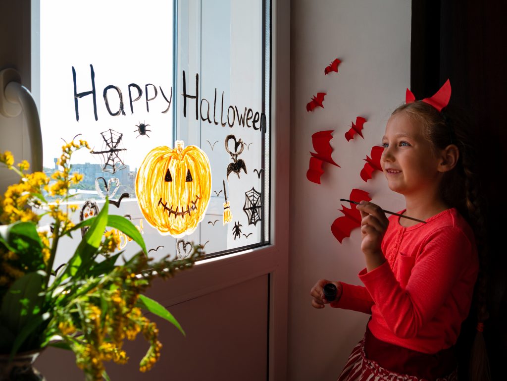 Child painting pumpkin on window preparing celebrate Halloween. Little kid draws decorates room interior with paper bats